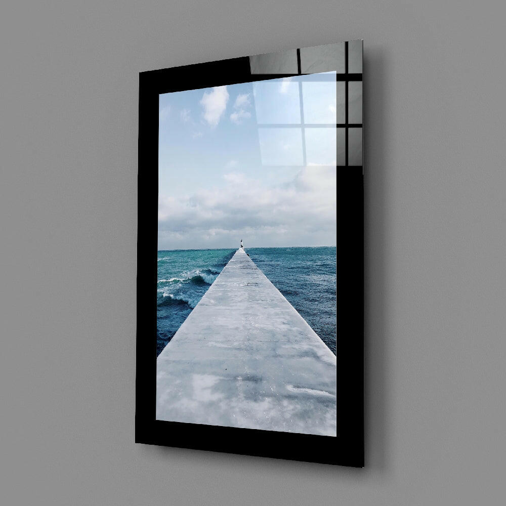 Black-framed glass wall art displaying a long white pier extending into turquoise ocean waves under cloudy sky.