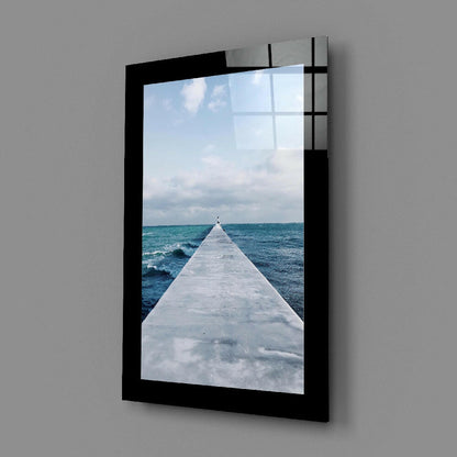 Black-framed glass wall art displaying a long white pier extending into turquoise ocean waves under cloudy sky.