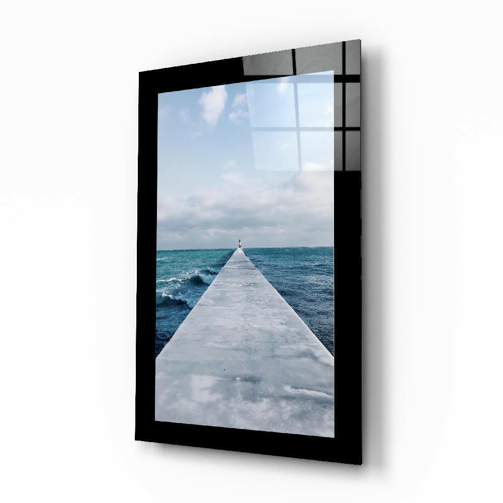 Black-framed glass wall art displaying a perspective view down a white concrete pier extending into turquoise ocean under cloudy sky.