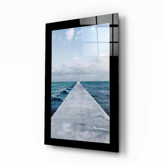 Black-framed glass wall art displaying a perspective view down a white concrete pier extending into turquoise ocean under cloudy sky.