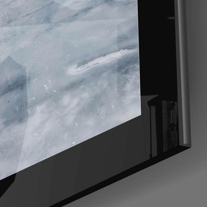 Glass wall art mounted on white wall displaying aerial landscape with snow-covered mountains and forested valleys in muted blue and white tones.