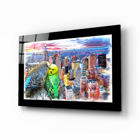 Black-framed glass wall art displaying two colorful budgies perched left of a vibrant cityscape skyline at sunset, mounted at an angle.