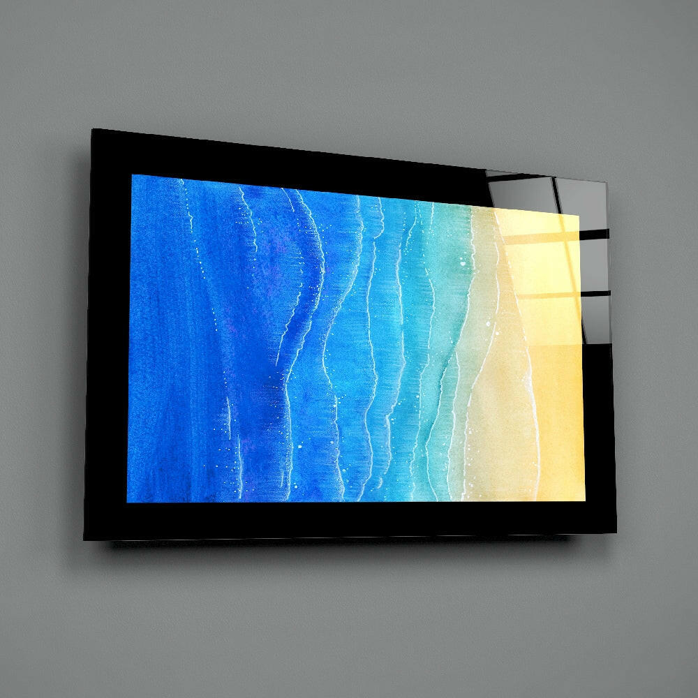 Black-framed glass wall art displaying aerial beach scene with blue ocean, turquoise water, and sandy shore, mounted on gray wall.