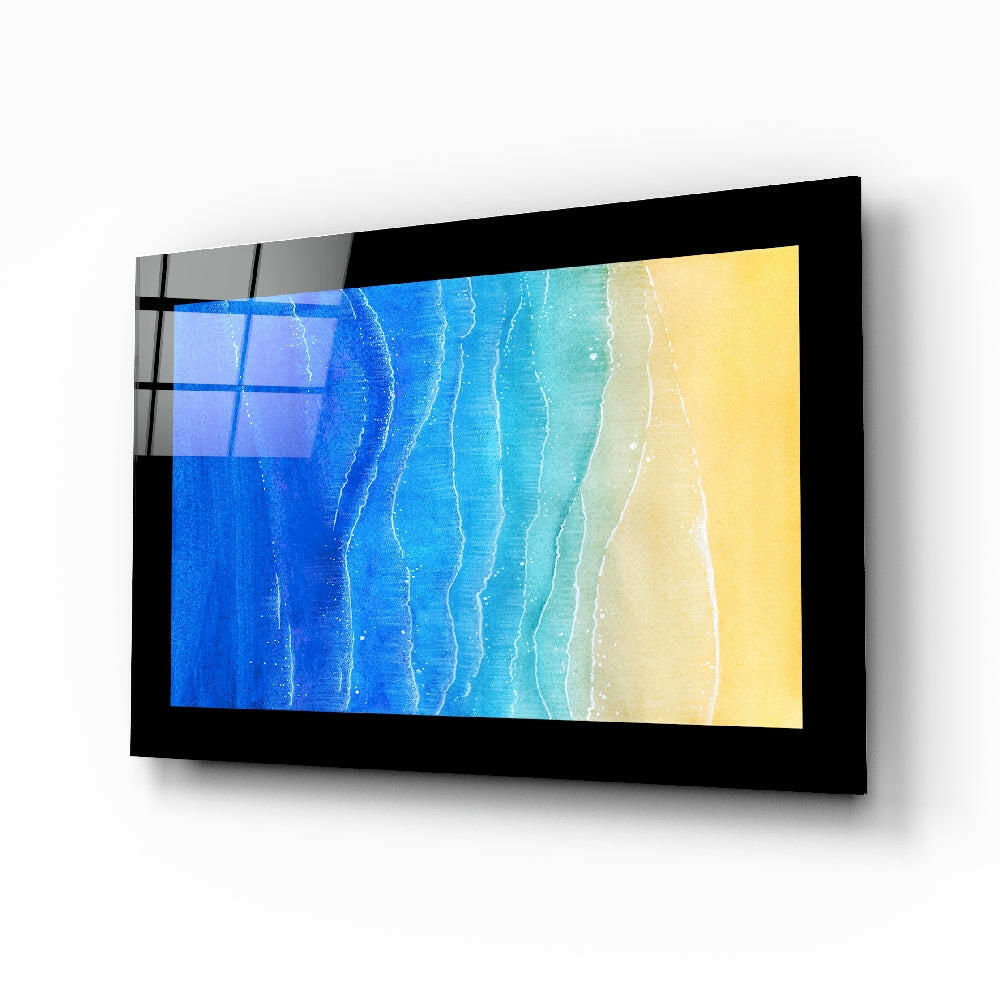 Black-framed glass wall art displaying aerial beach scene with blue ocean, turquoise waves, and yellow sand gradient, mounted at angle.