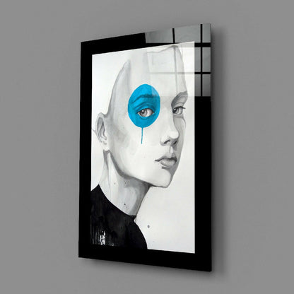 Black-framed glass wall art displaying a pencil-drawn face profile with a bright blue circular overlay on the eye, mounted at an angle against gray wall.