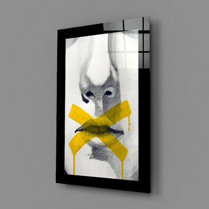 Black-framed glass wall art displaying sketched face with bright yellow X across mouth, angled perspective view.
