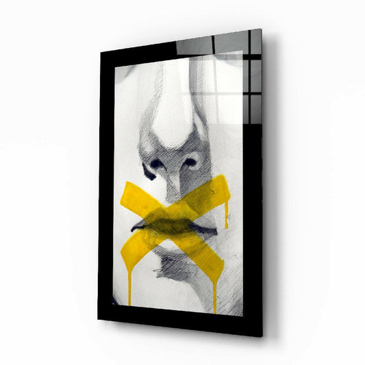 Framed glass wall art showing a sketched female face with a yellow X across the mouth, displayed at an angle against a white background.