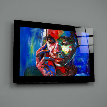 Mounted glass wall art displaying a portrait of a person with multicolored face paint in red, blue, green, and yellow tones against a blue background.