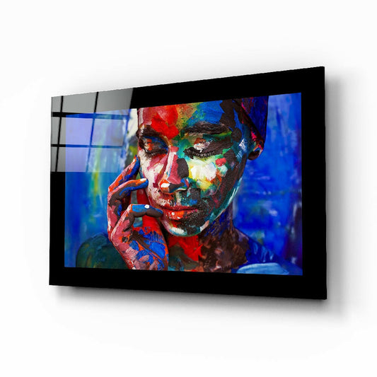 Black-framed glass wall art displaying a colorful portrait of a person with red, blue, yellow, and green paint on their face against a blue background.