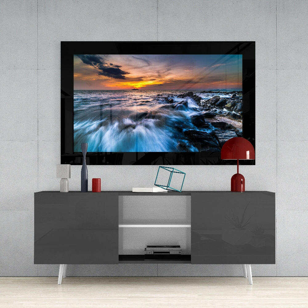 Framed coastal sunset photograph with crashing blue waves displayed above gray media console with white legs and red decorative accessories.