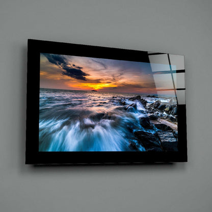 Black-framed glass wall art displaying a coastal sunset with turbulent waves and rocky shoreline mounted on gray wall.