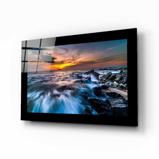 Black-framed glass wall art displaying a coastal sunset with rocky shoreline and crashing waves, mounted at an angle.