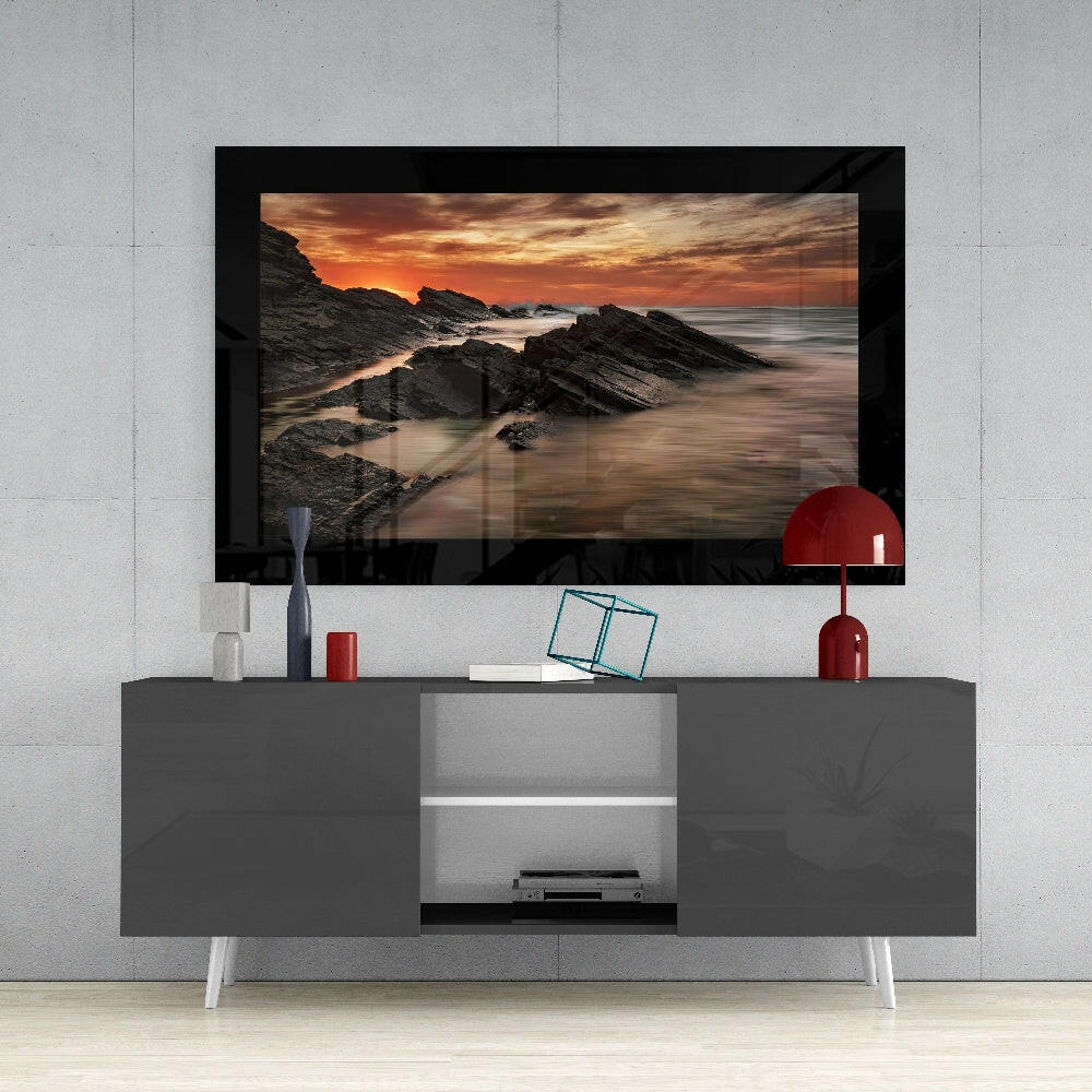 Framed glass wall art depicting a rocky coastline at sunset, displayed above a modern gray media console with white legs and red decorative accessories.