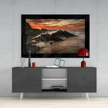 Framed glass wall art depicting a rocky coastline at sunset, displayed above a modern gray media console with white legs and red decorative accessories.
