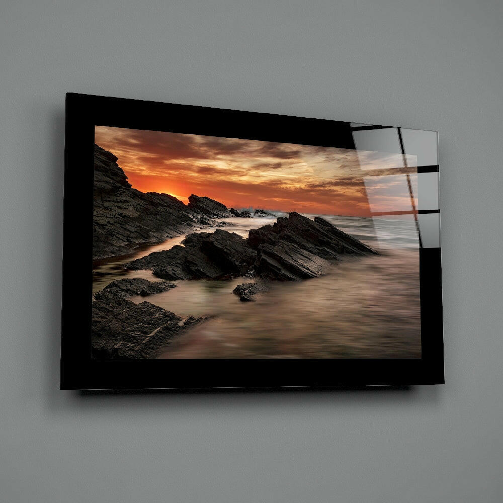 Black-framed glass wall art displaying a coastal sunset with rocky outcrops and golden sky, mounted on gray wall.