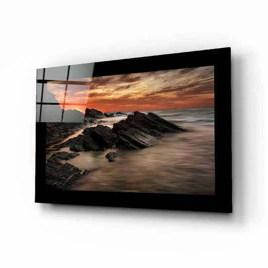 Black-framed glass wall art displaying a coastal sunset with rocky outcrops and golden sky, mounted at an angle.
