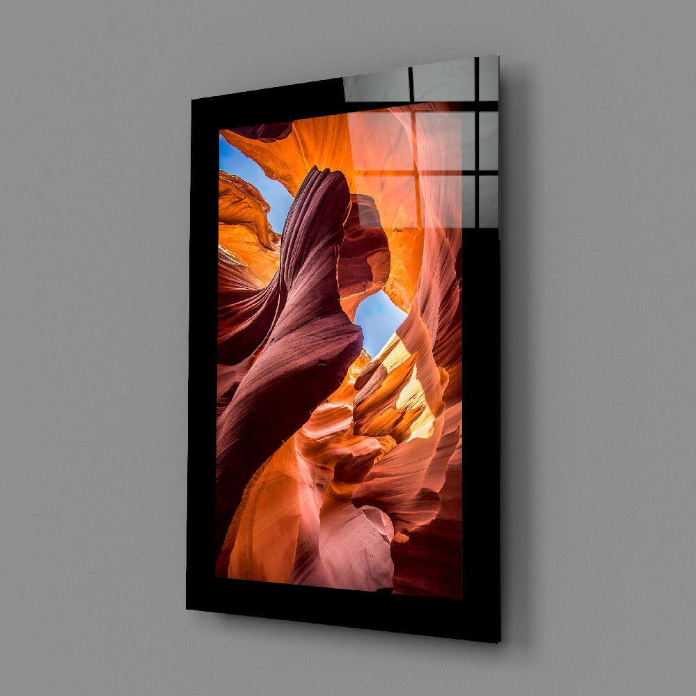 Black-framed glass wall art displaying a vibrant slot canyon photograph with orange, red, and golden rock formations and blue sky, mounted at an angle on gray wall.