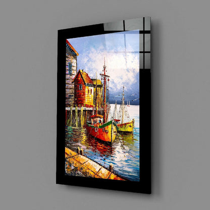 Black-framed glass wall art displaying a colorful painting of red and yellow fishing boats moored at a seaside village with stone buildings.