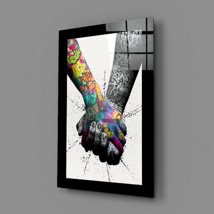 Black-framed glass wall art displaying two hands joining in vibrant graffiti colors against a white splatter background, angled three-quarter view.