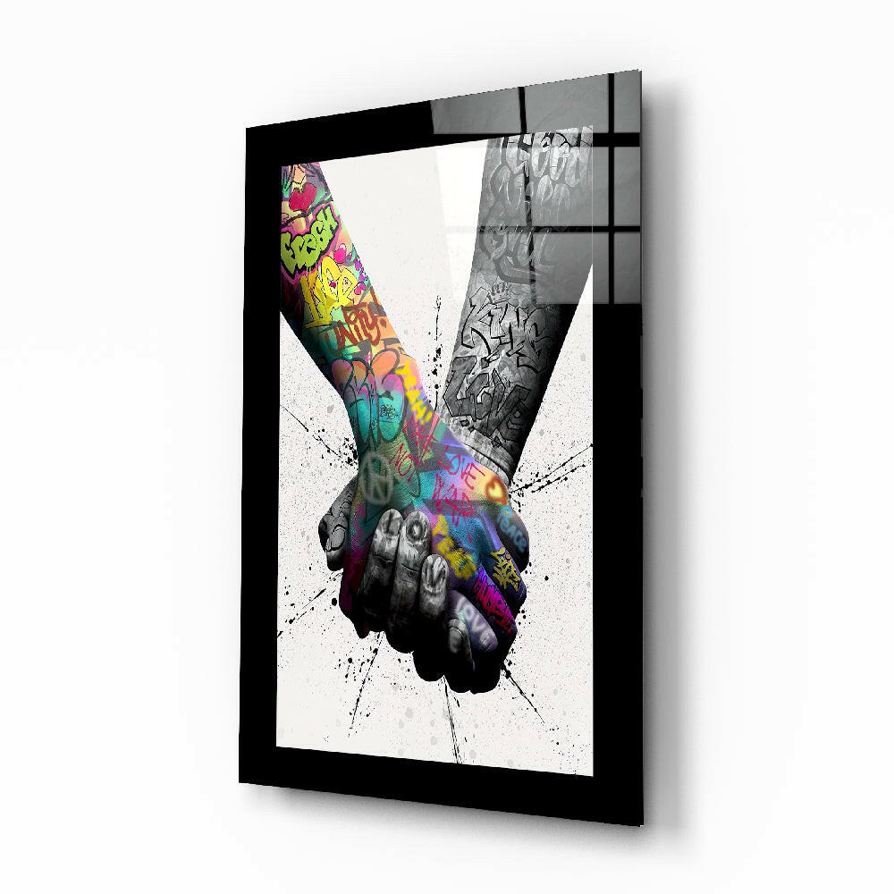 Black-framed glass wall art displaying two hands meeting with colorful graffiti-style artwork and splatter effects on white background.