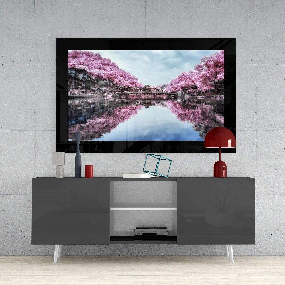 Framed glass wall art displaying pink cherry blossom trees reflected in water, mounted above gray media console with white legs.