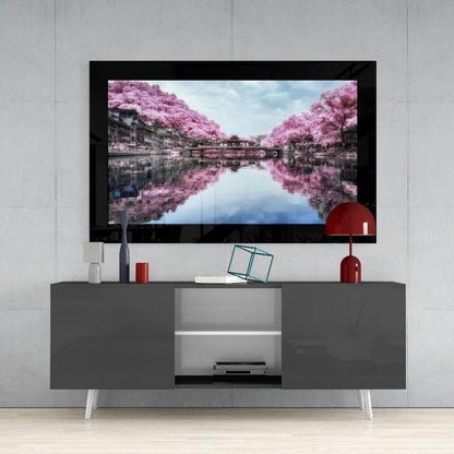 Framed glass wall art displaying pink cherry blossom trees reflected in water, mounted above gray media console with white legs.