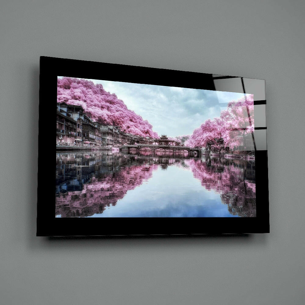 Black-framed glass wall art mounted on gray wall displaying pink cherry blossom trees reflected in still water with traditional architecture.