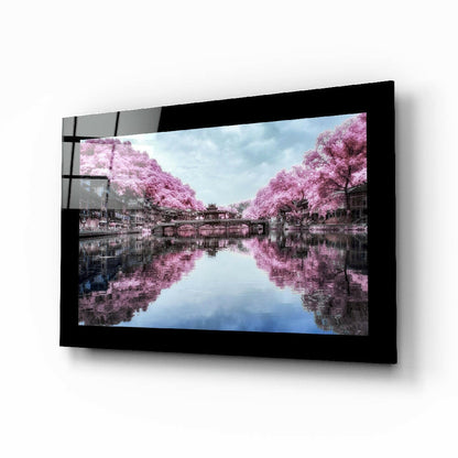Black-framed glass wall art displaying pink cherry blossom trees reflected in still water, mounted at an angle.