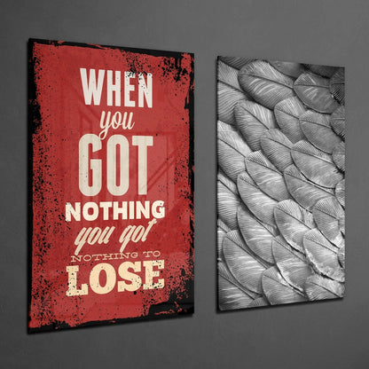 Two glass wall art panels mounted on gray wall: left panel red with distressed edges and cream text reading "When you Got Nothing you got Nothing to Lose"; right panel black and white feather pattern design.
