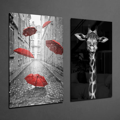 Two framed glass wall art panels displayed on gray wall: left panel shows rain-wet European street with red umbrellas in black and white; right panel shows black and white giraffe portrait.