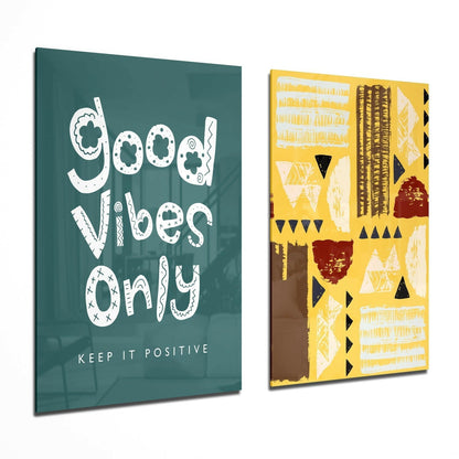 Two glass wall art panels displayed at an angle: one teal with white "Good Vibes Only" text, one yellow with geometric patterns in black, white, and red.