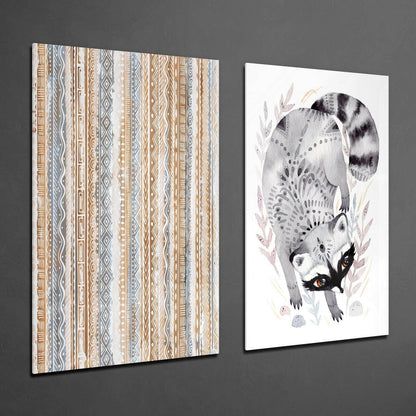 Two glass wall art panels displayed at an angle against gray wall: geometric striped pattern on left, illustrated raccoon with orange eyes and foliage on right.