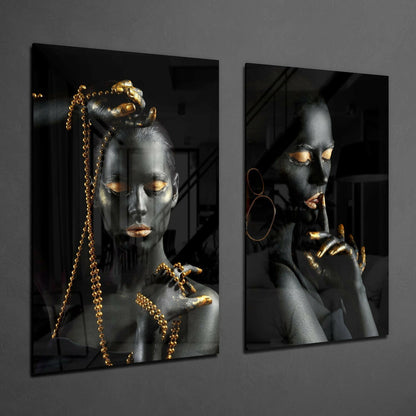 Two black and gold glass wall panels featuring painted female faces with gold leaf accents, gold chains, and hoop earrings against dark backgrounds.