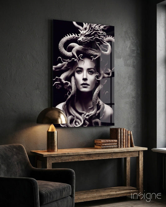 Monochromatic glass artwork depicting a woman's face with serpents and a dragon, mounted on a dark textured wall above a wooden desk.