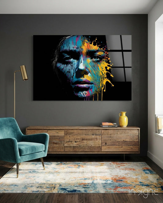 Colorful abstract portrait glass wall art featuring blue, yellow, and pink paint splashes on black background, displayed above wooden dresser in modern living room.