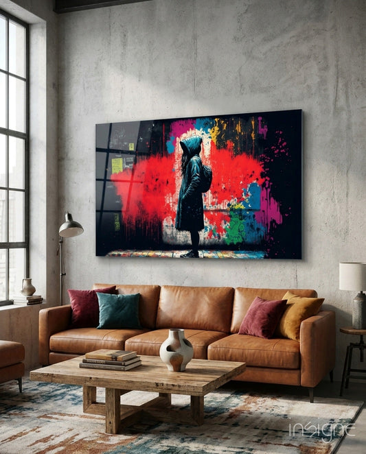 Glass wall art depicting a hooded figure standing before vibrant red, blue, and multicolored paint splashes on black background, displayed above a tan leather sofa.