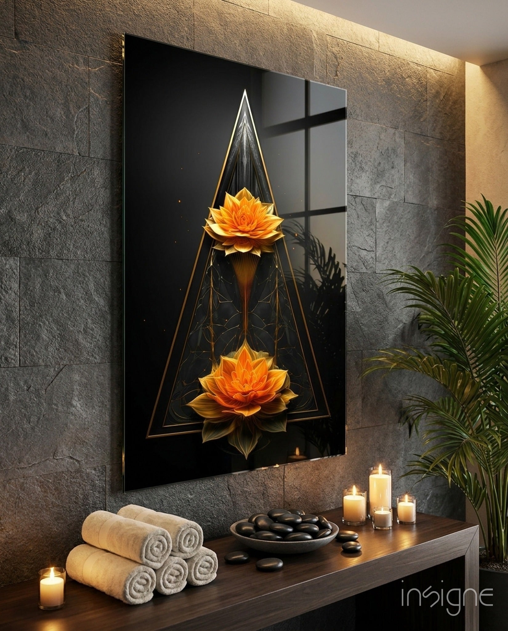 Black glass wall art featuring two golden lotus flowers in geometric triangular frame displayed above spa-themed wooden shelf with towels, stones, and candles.