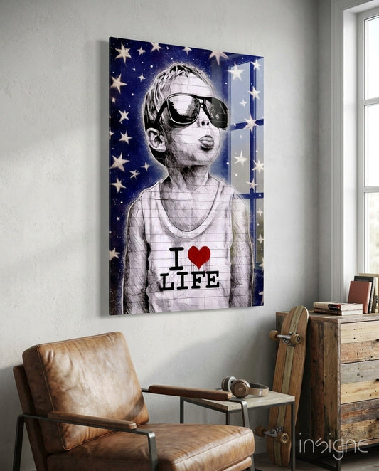 Glass wall art depicting a child wearing oversized sunglasses and "I ♥ LIFE" shirt against starry blue background, displayed in room setting above leather chair.