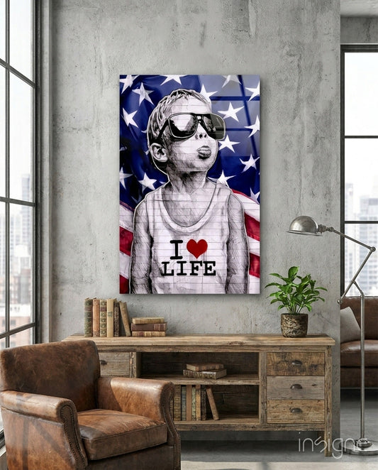 Glass wall art depicting a child wearing oversized sunglasses and "I ❤ LIFE" shirt against American flag background, displayed above wooden desk in industrial room.