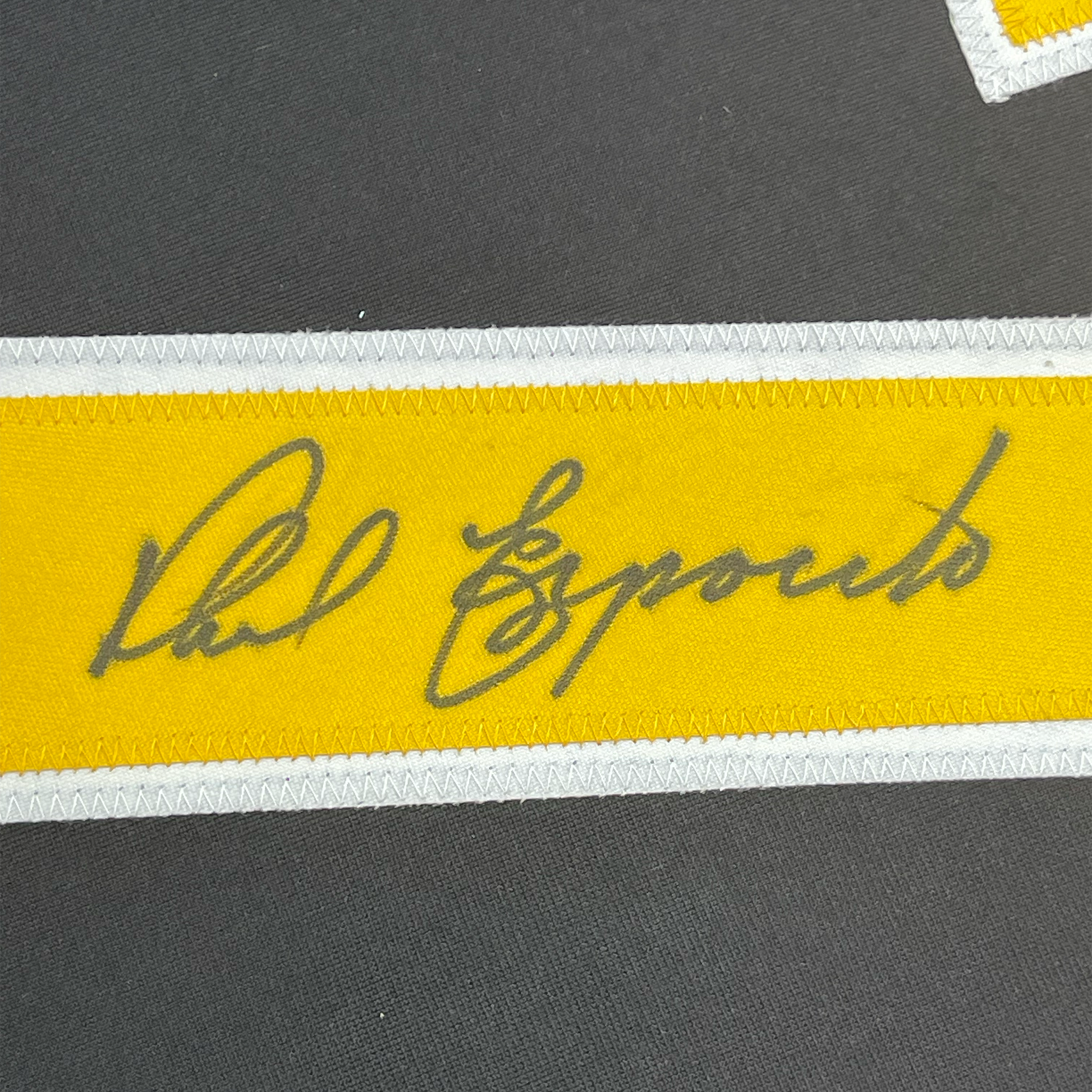 Close-up of signed yellow stripe on black jersey showing gray handwritten autograph reading "Phil Esposito" with white trim borders.