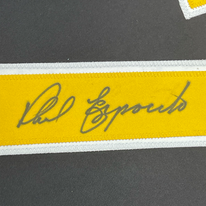 Close-up of signed yellow stripe on black jersey showing gray handwritten autograph reading "Phil Esposito" with white trim borders.