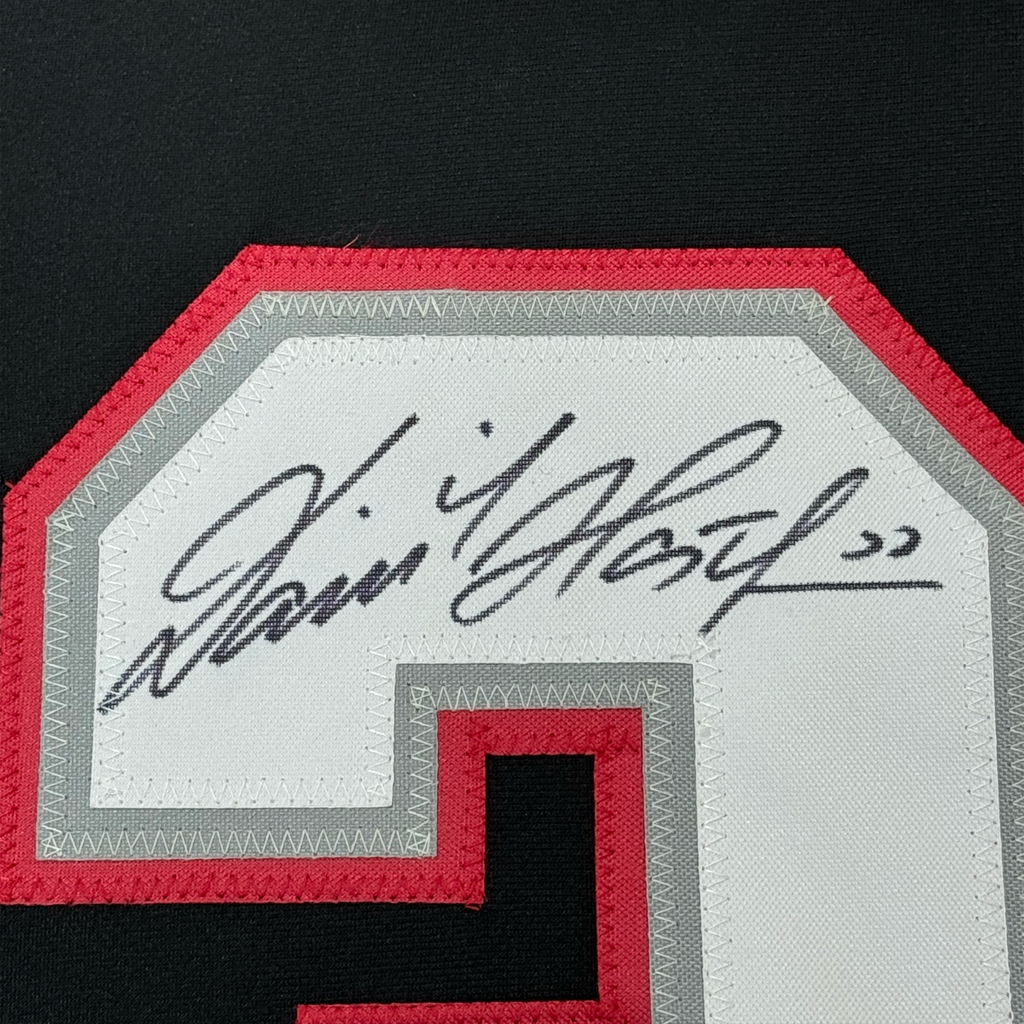 Close-up of handwritten signature on white jersey nameplate with red and gray stitched borders.