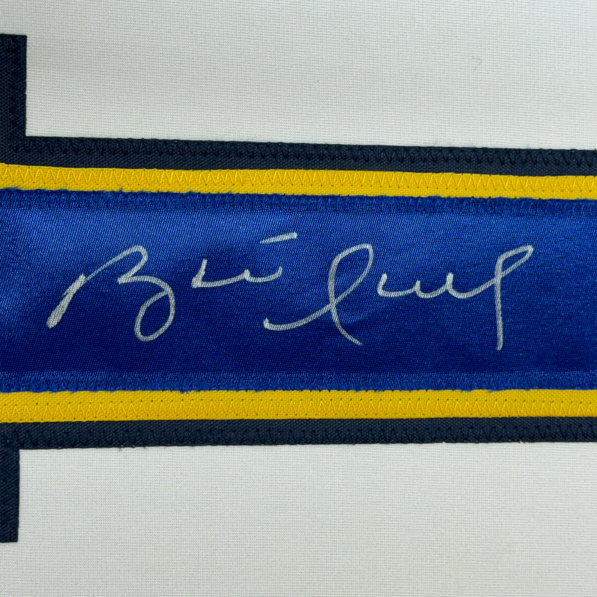 Close-up of blue hockey jersey stripe with handwritten signature in silver ink, yellow and black trim visible.