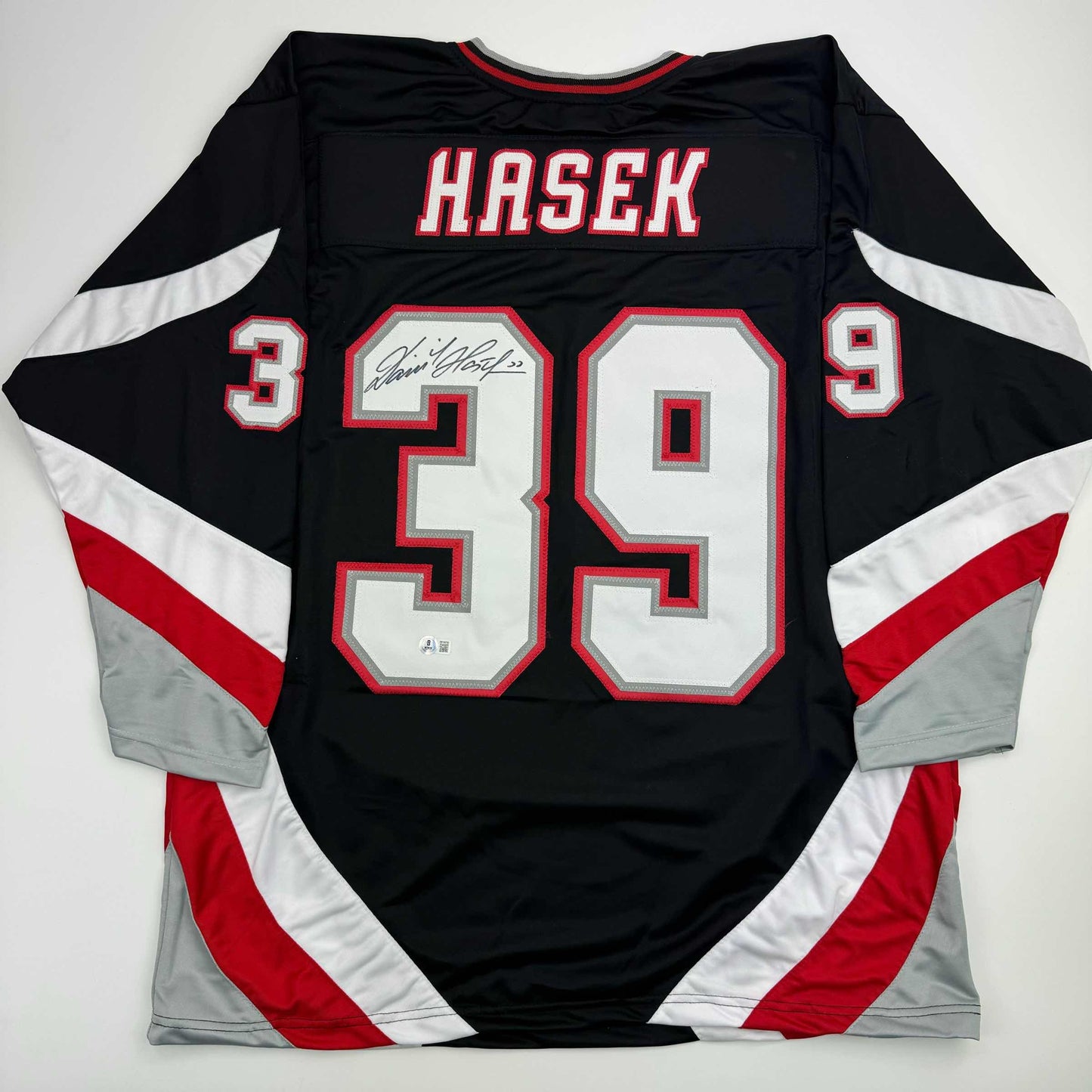 Back view of black, red, and white hockey jersey displaying "HASEK" and number 39 with autographed nameplate, laid flat.