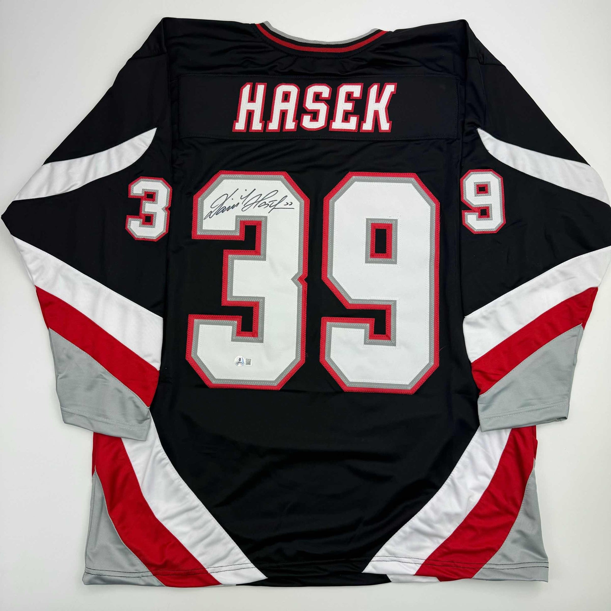 Back view of black, red, and white hockey jersey displaying "HASEK" and number 39 with autographed nameplate, laid flat.