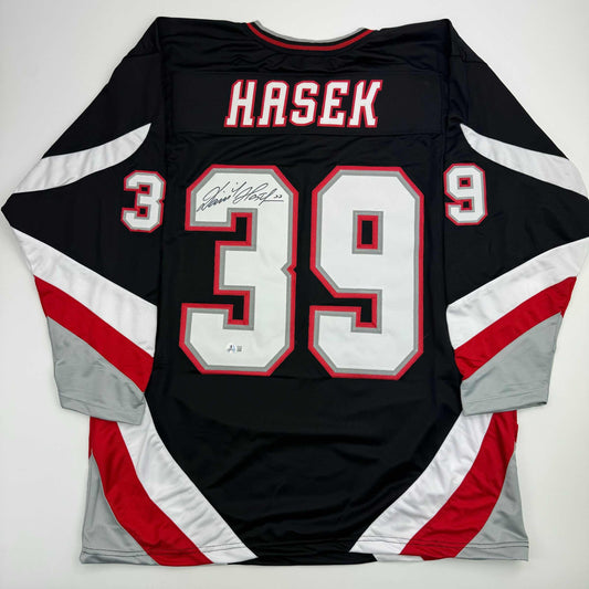 Back view of black, red, and white hockey jersey displaying "HASEK" and number 39 with autographed nameplate, laid flat.
