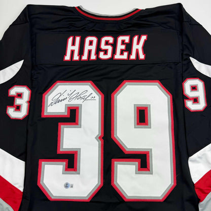 Back view of black hockey jersey with red and white number 39, "HASEK" nameplate, and autographed signature on white chest panel.