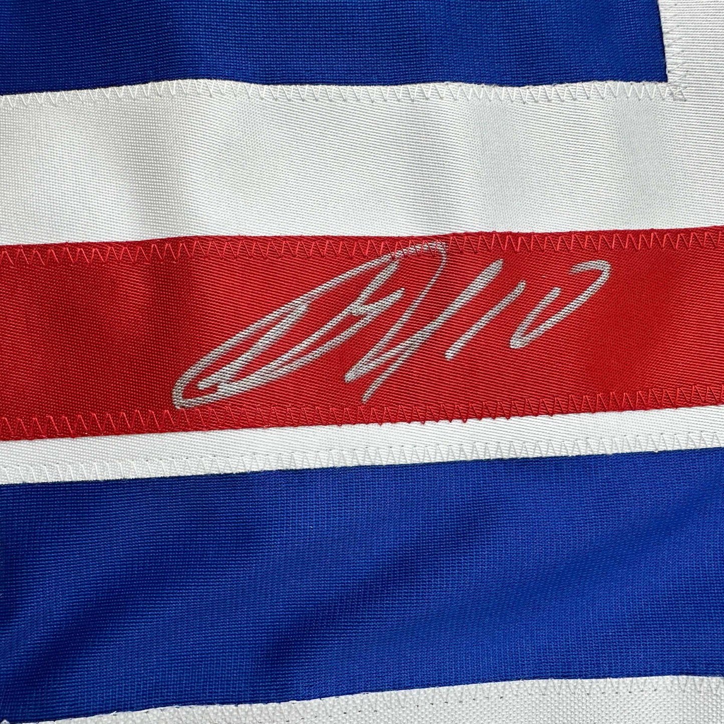 Close-up of signed red stripe on blue and white horizontally striped hockey jersey with gray cursive autograph.