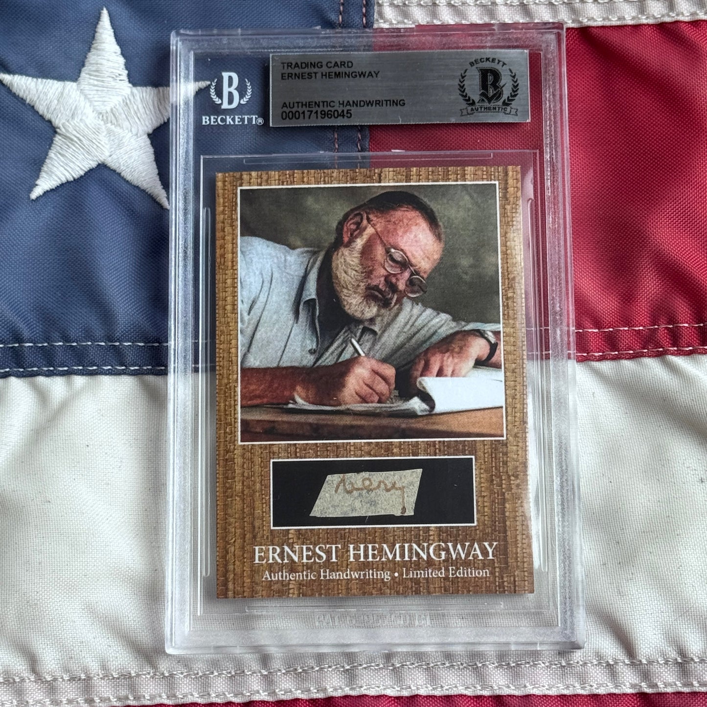 Beckett-authenticated Ernest Hemingway trading card featuring photograph of writer at desk with pen and authentic handwriting sample on wood-grain background, displayed on American flag.