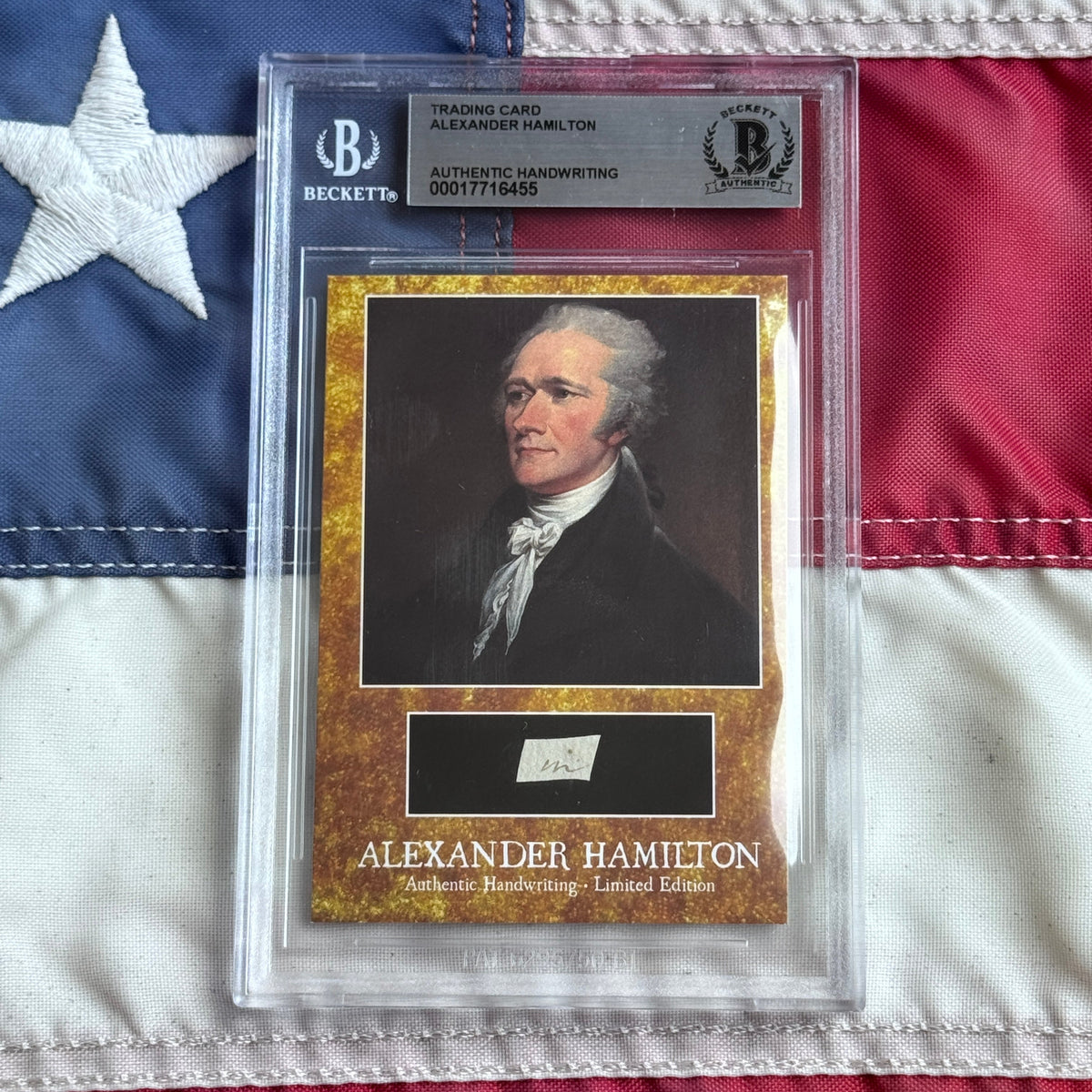 Beckett-authenticated trading card featuring Alexander Hamilton portrait with gold border and handwritten signature specimen, displayed against American flag background.